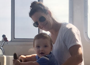 A mother and her baby son sit on the captain's chair at the wheel and look to the camera and smile.