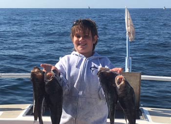 An adolescent boy smiles and holds up 2 sea bass in each hand.