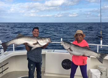 On a slightly overcast day on the water, a man and woman smile and each hold up a striped bass with two hands.