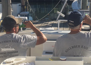 View from behind of Captain Steve and his father-in-law, Joe, both wearing light gray November Rain t-shirts and drinking.