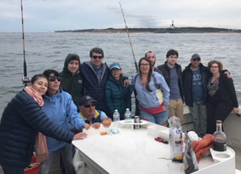 A family of eleven stand on the boat and smile for the camera with the lighthouse behind them in the distance.
