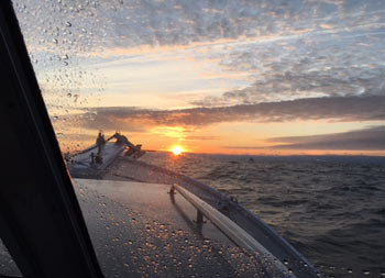 Close up view from inside of the boat's cabin through a rainy window of the sun on the horizon in the distance.