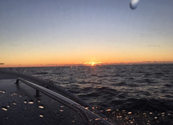 View from inside the cockpit looking out the window at the orange/yellow/blue sky while the sun sets on the horizon of the dark blue water.