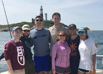 A group of 7 men and women smile for the camera as they stand on the boat with the lighthouse behind them.