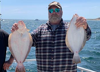 A man with a beard, baseball cap, sunglasses and flannel shirt, holds up 2 fluke, one in each hand.