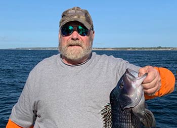 A man with a beard, baseball cap and sunglasses holds up a sea bass.