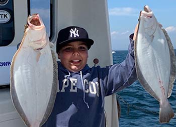 An adolescent boy smiles as he holds up the two fluke he caught.