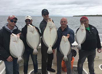 A group of 5 men each smile and hold up the fluke they caught.