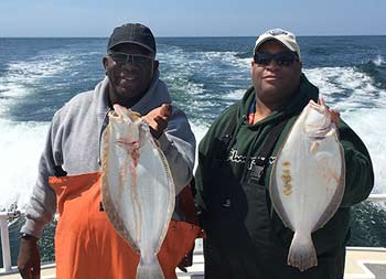 Two men wearing sunglasses on a sunny day each hold up fluke.