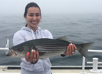 A woman with brown hair smiles and holds up the striped bass she caught.