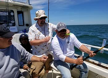 A man jokingly helps out his friend reel in a fish as other look on and laugh.