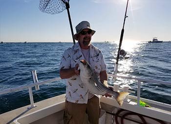 With the sun reflecting brightly on the water, a man wearing a hat holds up the fish he caught.