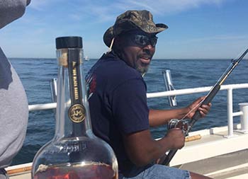 A man sitting in the background with rod in hand, looks back towards the camera and smiles as we get a glimpse of a bottle of Jameson whiskey in the foreground.