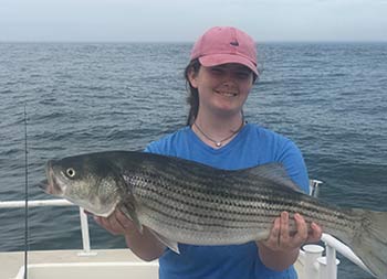 On an overcast day, a teenage smiles and holds up the striped bass she caught .