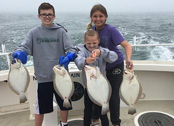 A teenage boy and girl each hold up the fish they caught. The younger girl has a face of determination, looking at her catch and using all her strength to hold it up .