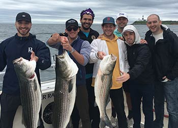 With the lighthouse behind them in the distance, seven men hold up the striped bass they caught and smile for the camera.
