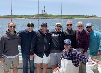 Seven men, including three generations, line up and smile for the camera with the lighthouse displayed in the background and Captain Steve in front kneeling for the photo.