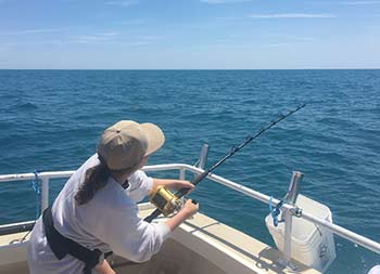 View from behind of a woman using her strength to reel in a shark that's in the water out of sight.