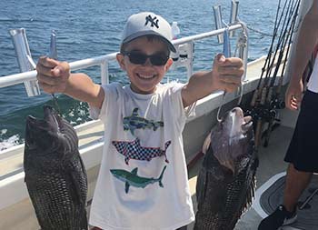 A young boy wearing sunglasses holds up a sea bass in each hand.