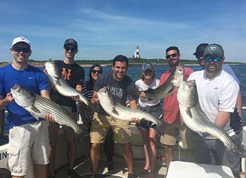 With the lighthouse in the distance, a group of 8 men and women hold up the striped bass they caught.