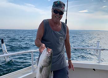 With the clouds in the blue sky above, a man with sunglasses smiles big as he holds up the striped bass he caught.