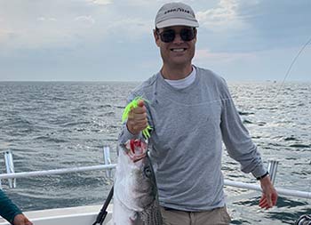 With the sun setting in the background, a man smiles and holds up the striped bass he caught.