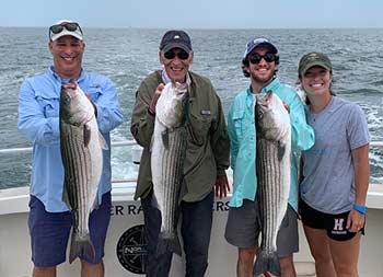 A man with his father and two young adult children, a boy and girl, each hold up a striped bass and smile for the camera.