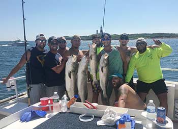 With the lighthouse in the distance behind them, a group of 9 guys smile and hold up the fish they caught.