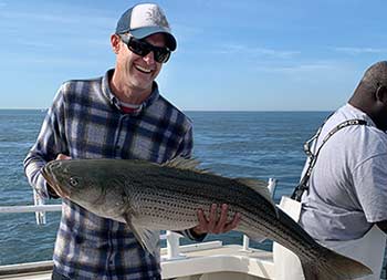 With the sunny blue sky and ocean behind him, a man wearing sunglasses, smiles and holds up a striped bass
