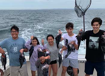 A mix of six young, adolescent and teenage boys and girls smile and hold up the sea bass they caught.