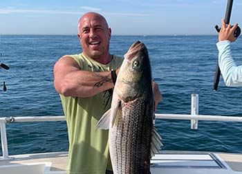 With the blue sky and ocean behind him, a man wearing sunglasses holds up the striped bass he caught and smiles.