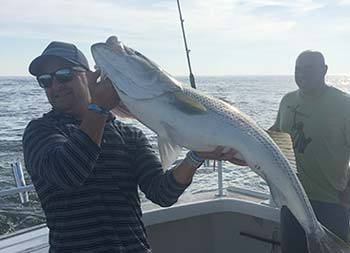 With the sun behind them, a man holds up the striped bass he caught and another man behind him smiles.