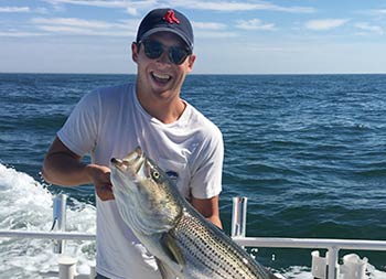 With the blue sky and ocean behind him, a man wearing a baseball cap and sunglasses holds up the striped bass he caught and smiles.