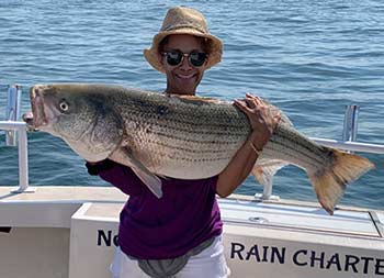 On a sunny day, a woman wearing a sun hat and sunglasses holds up a striped bass in her arms.
