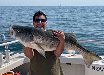 On a sunny day, a young man wearing sunglasses smiles holds up a striped bass in his arms.