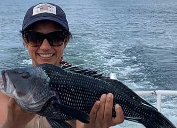 A woman holds up the sea bass she caught and smiles.