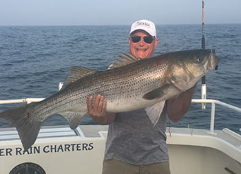 A man holds up a striped bass with both hands as he smiles.