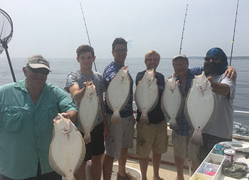 On an overcast grayish day, a group of six hold up the fluke they caught and smile for the camera.