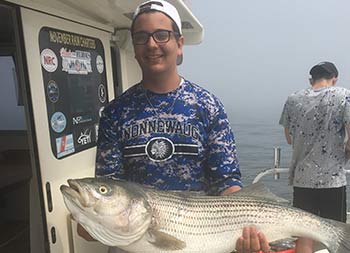 With an overcast grayish sky, a young man holds up a striped bass with both is arms .