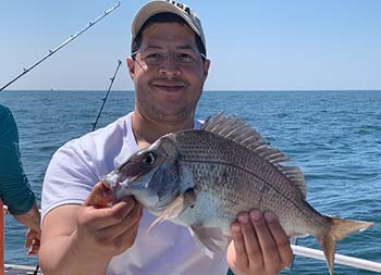 With the blue sky and ocean behind him, a man smiles for a close up holding porgy(scup).