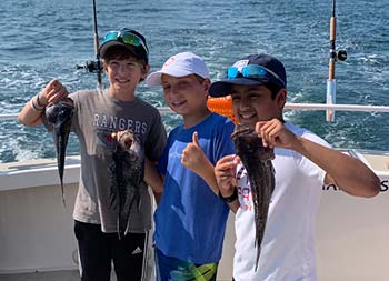 Three young boys each hold up sea bass and give thumbs up/peace sign.