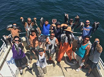 View from atop the boat of thirteen men, including first mate, Gil, looing up at the camera, each holding 2 sea bass.