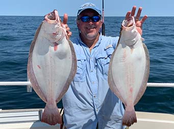 With the blue sunny sky and ocean behind him, a man wearing sunglasses smiles and holds up the fluke he caught.
