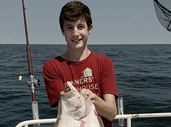 A young man wearing a dark red tshirt holds up the fluke he caught .