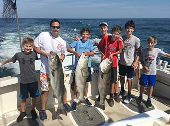 Two men and four adolescent boys hold up 3 striped bass and smile for the camera.