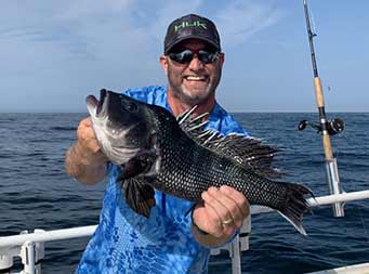 A man wearing a sunglasses smiles big and holds up the sea bass he caught with both hands showing it to the camera.