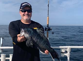 Another man wearing a sunglasses also smiles big and holds up the sea bass he caught with both hands showing it to the camera.