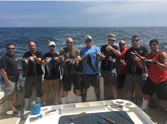 With the ocean behind them, nine men standing together each hold up 2 sea bass.