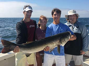 A father, his daughter and two sons stand together as the father and a son hold up a striped bass.