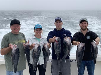 With the dense fog and wake of the boat behind them, three men and one woman smile and each hold up 2 sea bass.